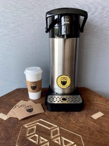 A coffee maker on a table with a logo etched into it. On the left, coffee supplies are laid out with the Morning Owl logo, and the stainless steel coffee maker has a bright yellow Morning Owl sticker on it. 