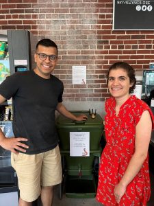 Two innovators stand smiling in front of what's known as The Box of Life - a composting solutions that uses worm power to transform food waste into powerful soil. 