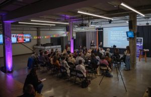 A view from above a group sitting in front of a presentation on a screen. The pillars in the room are glowing purple. 