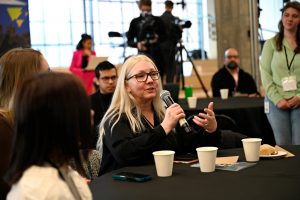  A person holds a microphone while seated at a table, in discussion at an in-person event. 