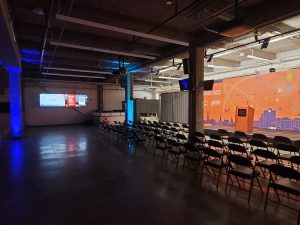 An open space event room is shown that is dark, with concrete pillars that are accented in purple and blue. 