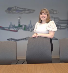 A person stands in from of a mural of a cityscape on a wall - with their arms resting on the top of a chair that's pulled up a boardroom table. 