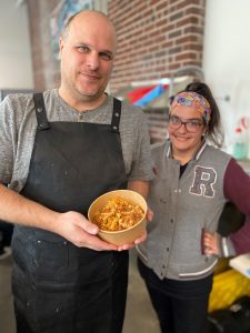 Two food workers stand side by side in a cafe showcasing a bowl of food they have prepared, smiling. 