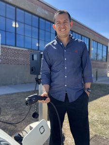 JPlug Founder Jorge Pantoja standing smiling, holding an electric car charging unit at a car. 