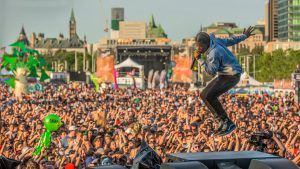 A performer is singing and leaping on stage in front of a large crowd at an outdoor festival in Ottawa during the daytime.