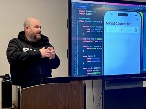 A presenter is speaking in beside a display screen showing code and a mobile device. 