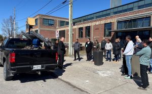A group gathers outside for a technology demonstration. A man stands in front of a parked black pickup truck.