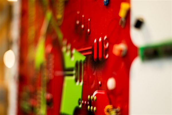 Close-up of a vibrant art installation at Bayview Yards featuring a bright red panel with protruding pegs, pins, and small colorful components, including a green geometric shape, shown in shallow focus with blurred elements in the background.